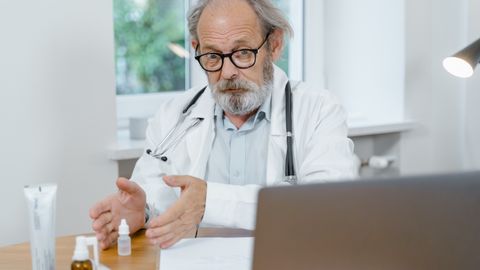 An elderly doctor in a white coat conducting an online medical consultation, focusing attentively.