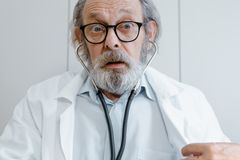 Elderly doctor with beard and glasses using a stethoscope in a medical setting.