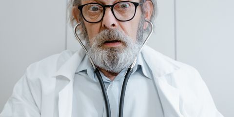 Elderly doctor with beard and glasses using a stethoscope in a medical setting.