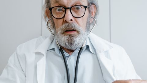 Elderly doctor with beard and glasses using a stethoscope in a medical setting.