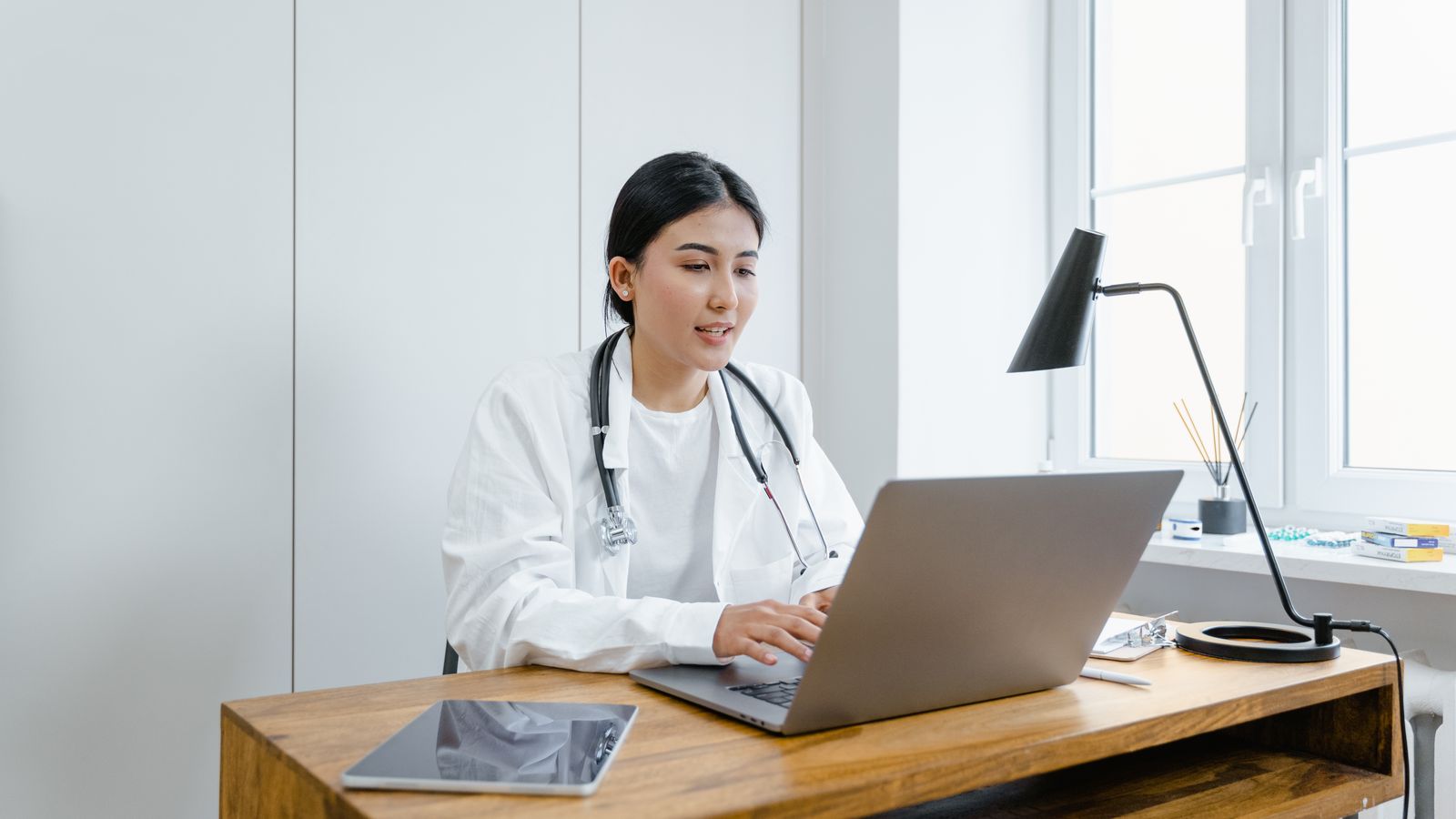 A female doctor in a white coat uses a laptop for an online consultation from her office.
