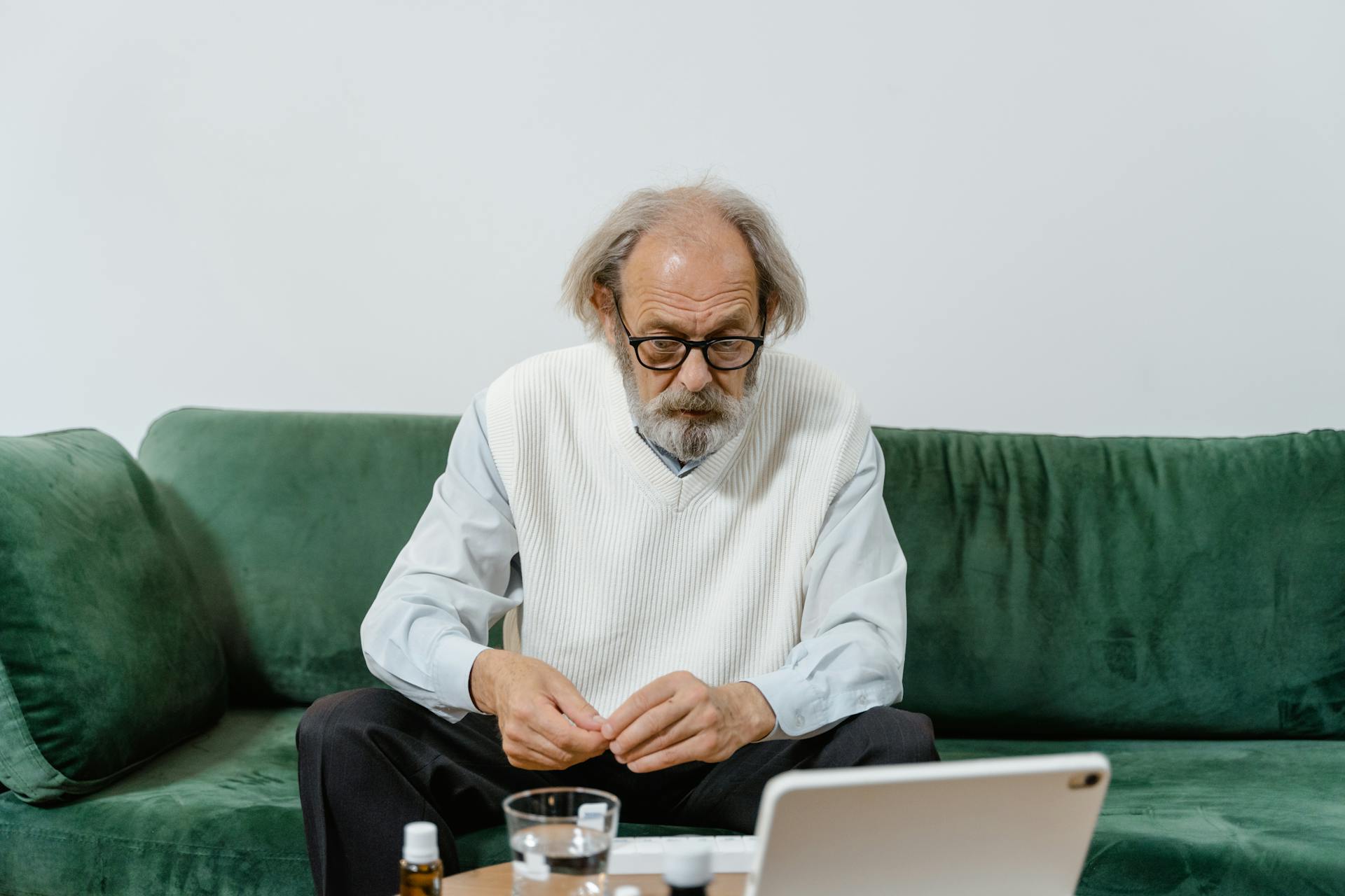 Senior man sitting on green sofa, focusing on a tablet, possibly for telemedicine or social interactions.