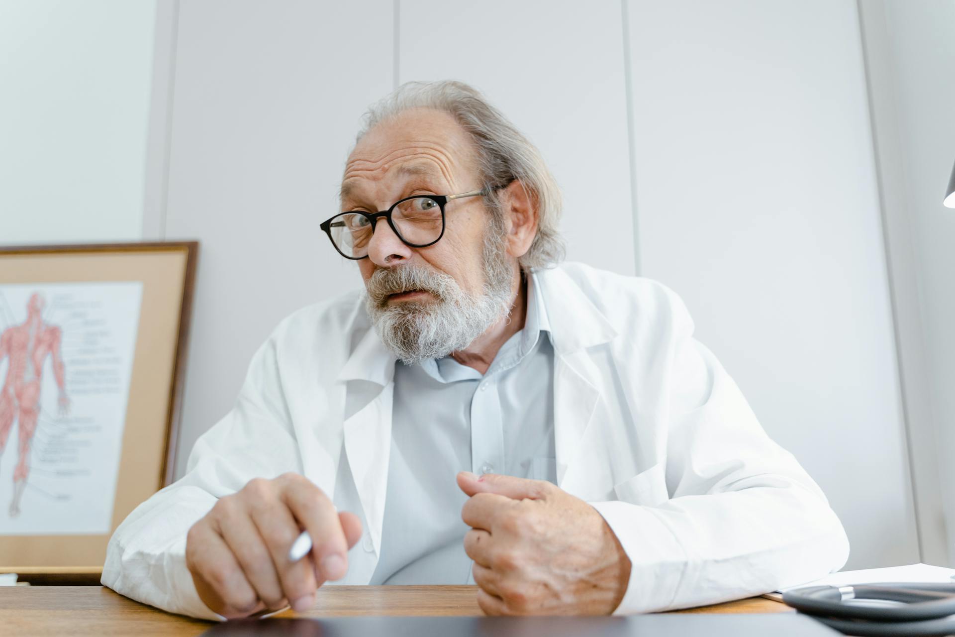 Elderly male doctor in office, wearing glasses, ready for telemedicine session.