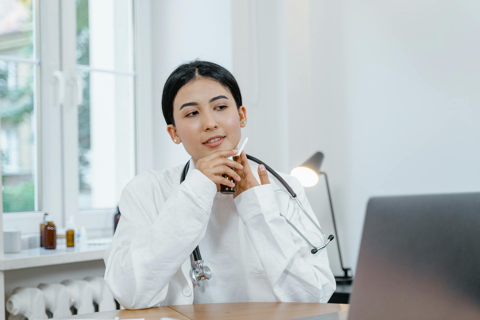 Asian female doctor using a smartphone for a video consultation, seated in a bright office setting.