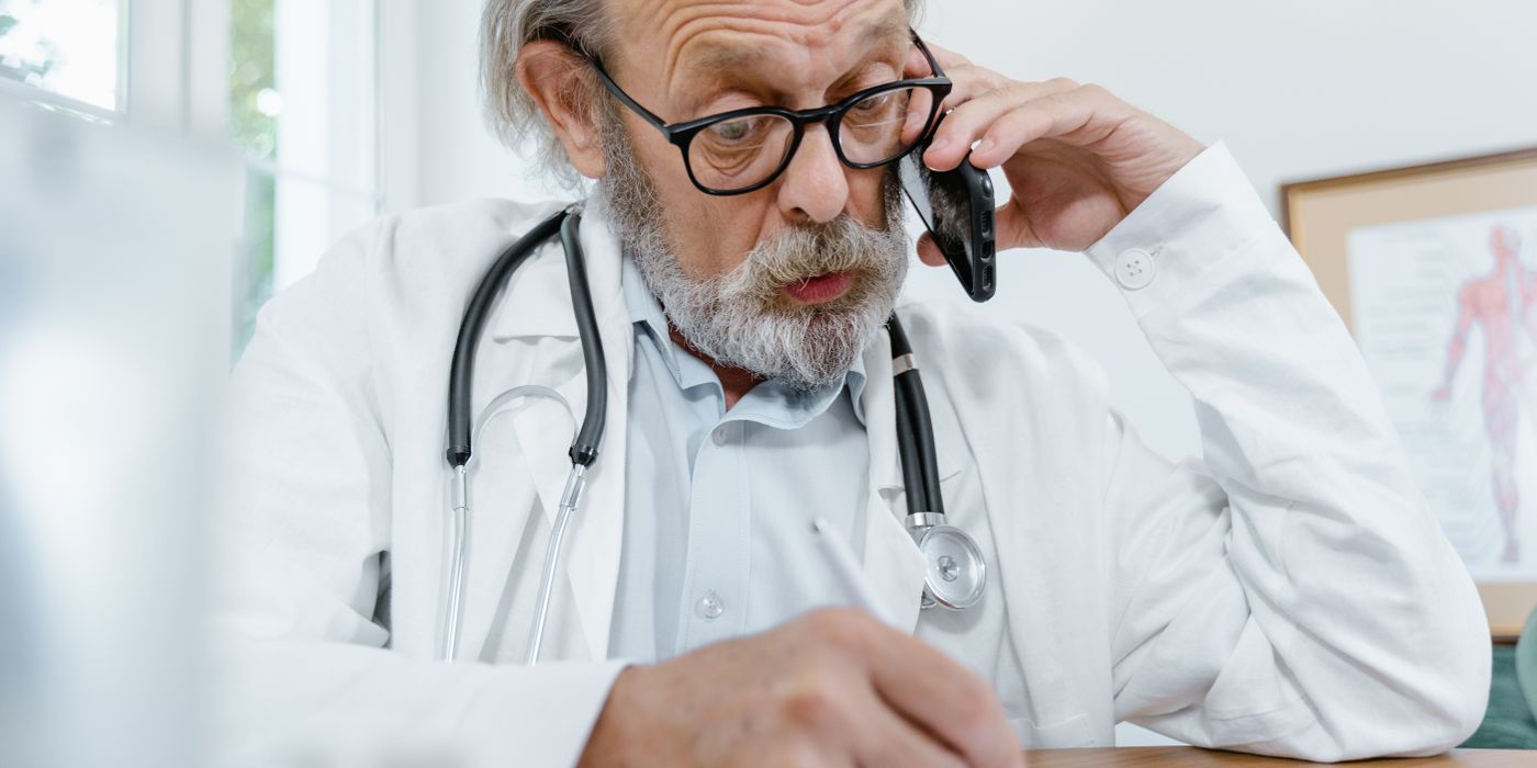 Elderly doctor in office on phone call with patient, writing notes.