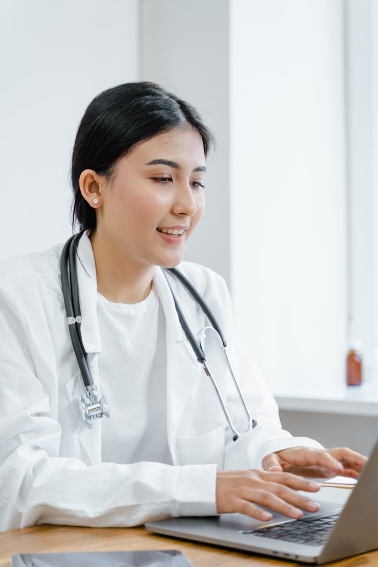 A Woman In White Lab Coat Smiling While Typing On Laptop