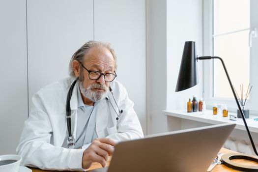 Senior male doctor in lab coat using laptop for video call in clinic office.