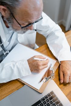A doctor writing on a clipboard, working at a wooden desk with a laptop.