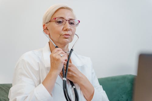 Free Elderly female doctor using a stethoscope during a virtual consultation in a medical office. Stock Photo