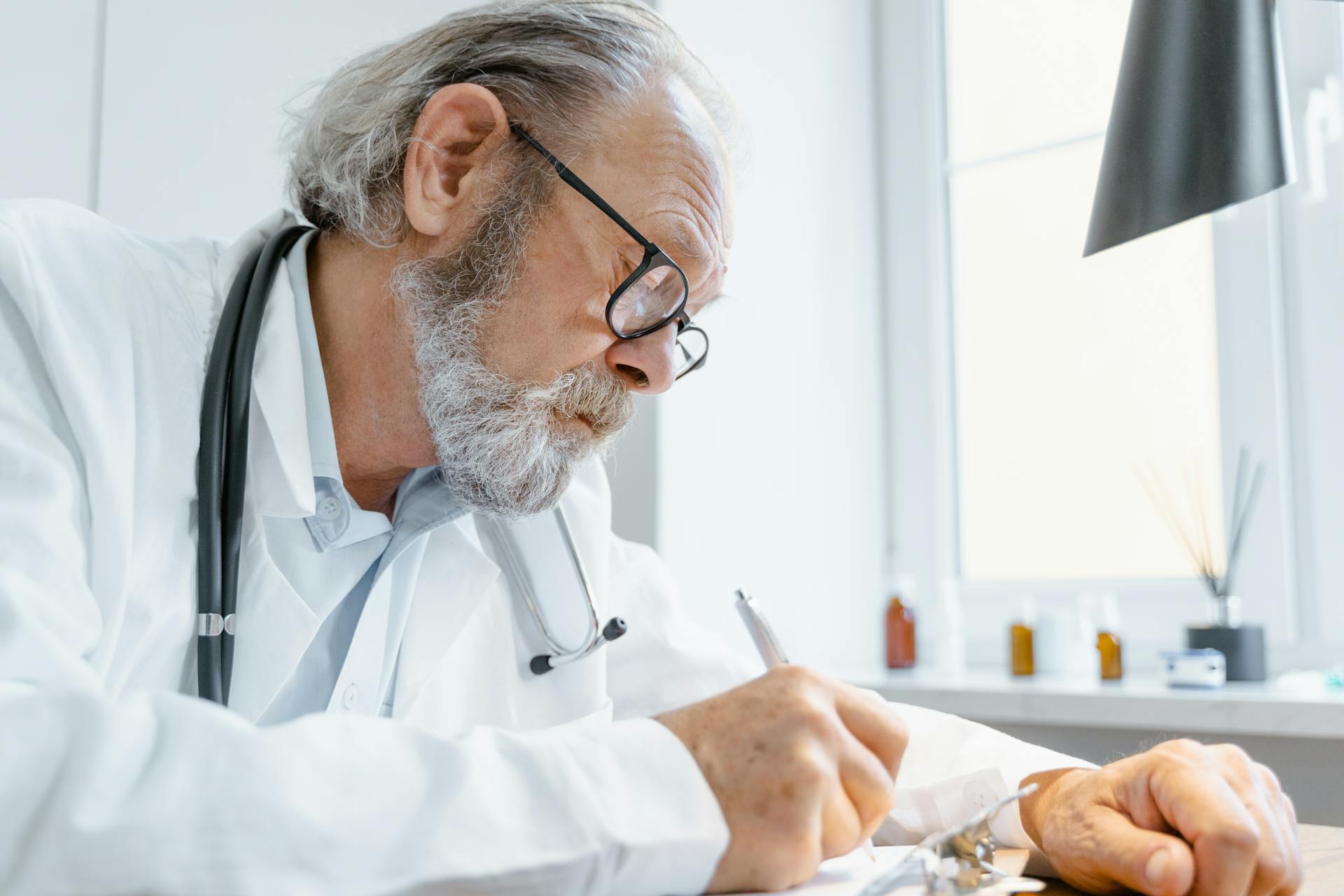 Elderly male doctor writing notes in a bright medical office.