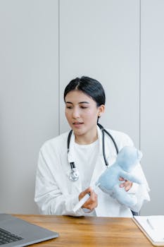 A female pediatrician with a stethoscope is in a clinic, holding a syringe and a stuffed toy bear.