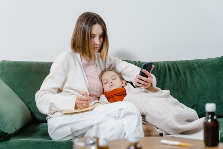 Woman Using Phone With Sick Child Sleeping In Her Arms