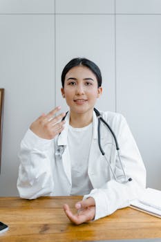 A professional young female doctor in a white coat, smiling and gesturing indoors.