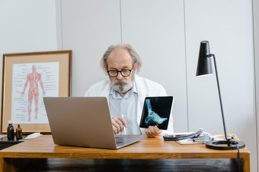 Elderly male doctor examines X-ray on tablet while working at his office desk with a laptop.