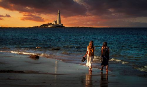Free Two women strolling along a beach at sunset with a lighthouse in the background, creating a serene seascape. Stock Photo