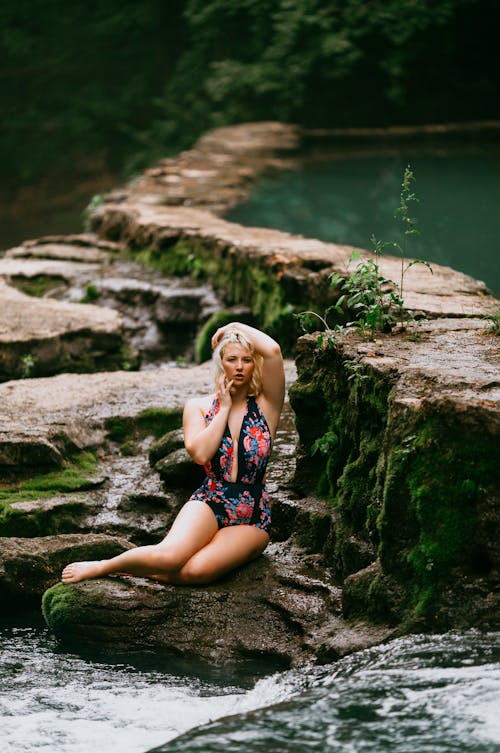 Free A Woman in Floral Swimsuit Sitting on a Mossy Rocks Near the Stream while Posing at the Camera Stock Photo