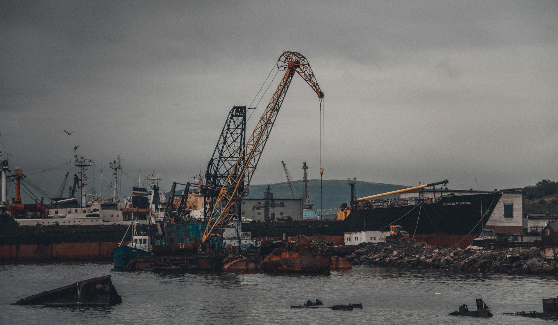 Moody industrial dock scene with cranes and cargo ships under a cloudy sky.