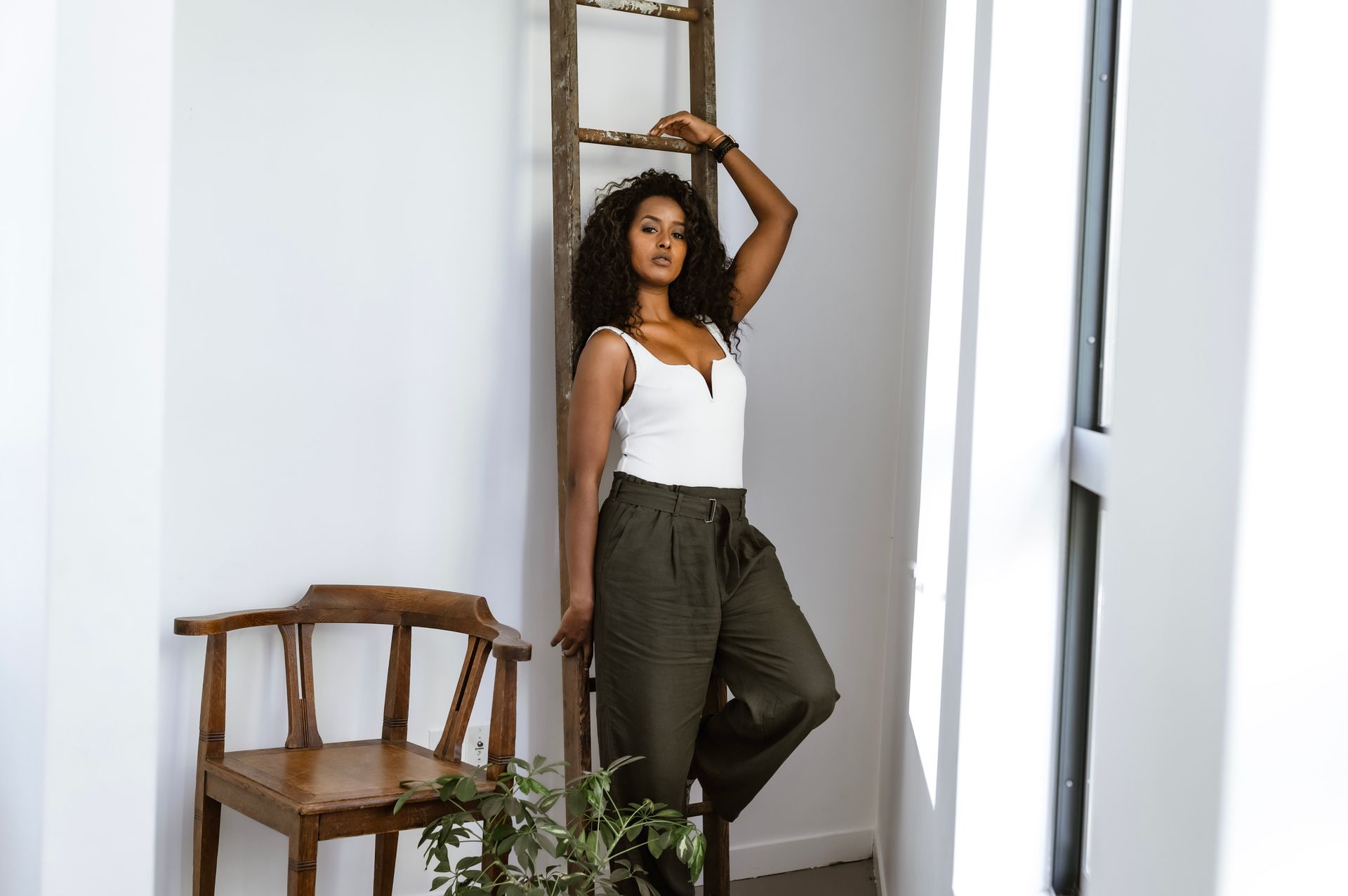Chic woman in white top and green pants posing stylishly indoors with ladder and chair decor.