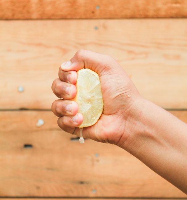 A hand tightly squeezes a lemon, releasing fresh juice against a wooden backdrop.