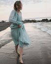 Full body of barefoot female in long wavy dress walking along sandy beach near sea while smiling in summer evening