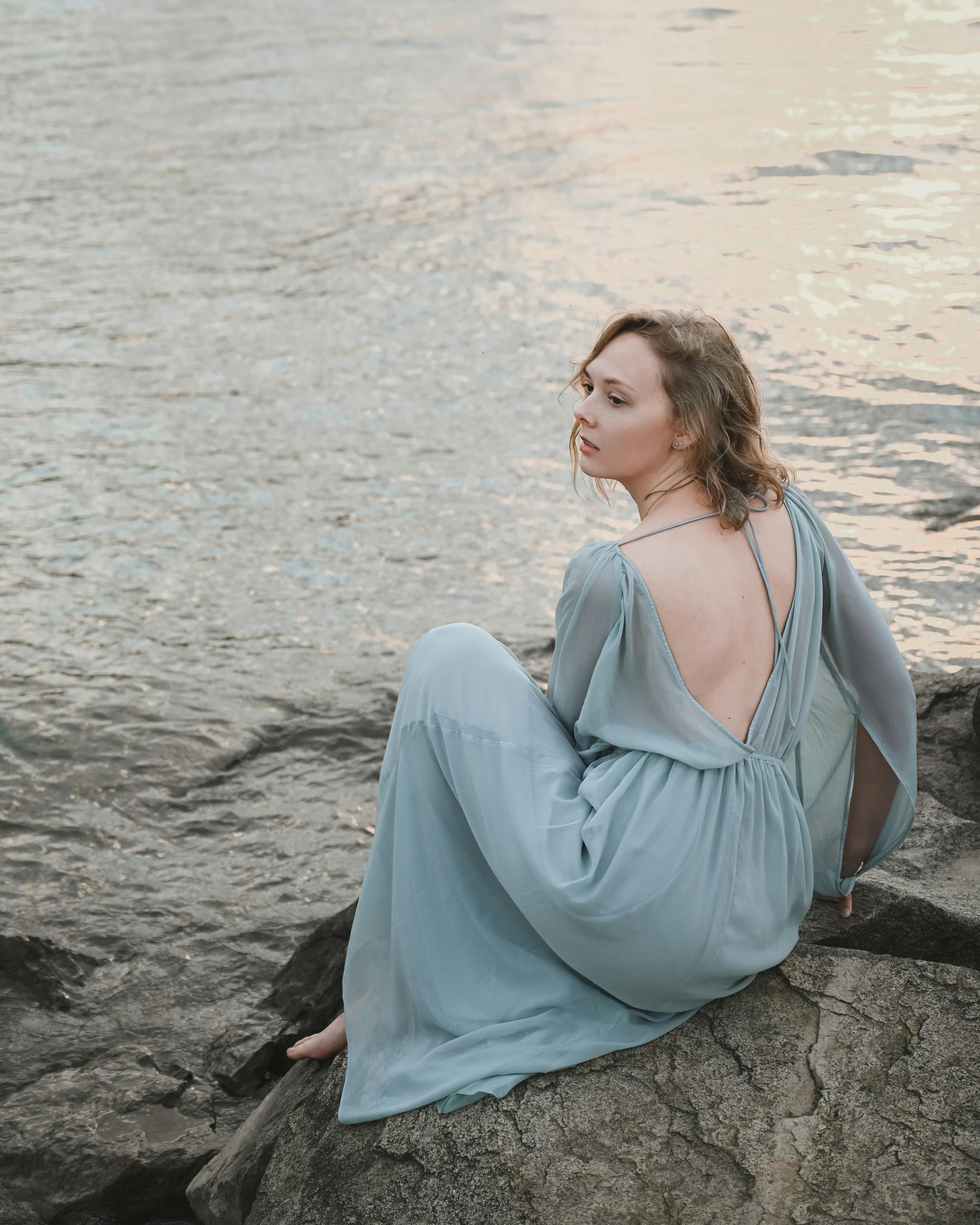 一名身穿飘逸蓝色连衣裙的女子优雅地坐在海边的岩石上，凝视着水面