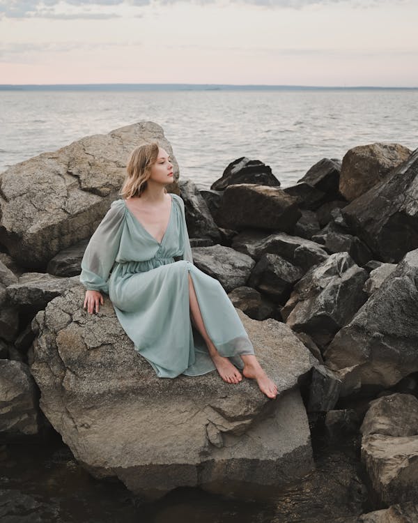 Full body of barefoot calm female wearing blue dress sitting on rocky coast of rippling sea and looking away dreamily on cloudy day