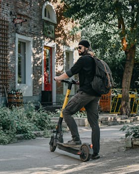 Full body of unrecognizable bearded hipster male in black outfit and hat with black sunglasses and backpack standing on electric kick scooter near brick building and looking away in sunny day