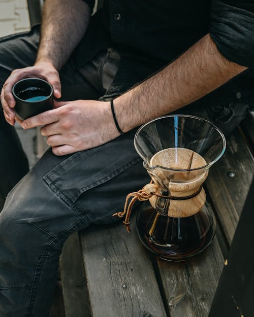 From above of crop unrecognizable male in black outfit sitting on wooden bench with coffee pot and holding cup of coffee