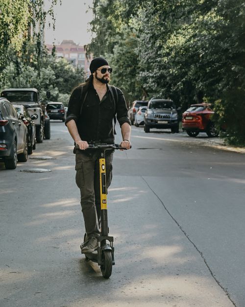 Free A stylish man with a bandana and sunglasses rides an electric scooter down a leafy urban street. Stock Photo