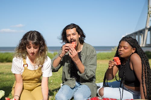 People Sitting on a Grassy Ground while Eating Sliced of Fruit