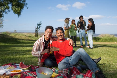 Men Sitting on a Picnic Blanket while Holding a Bottle of Alcoholic Drinks Near Group of People Standing while Having a Conversation