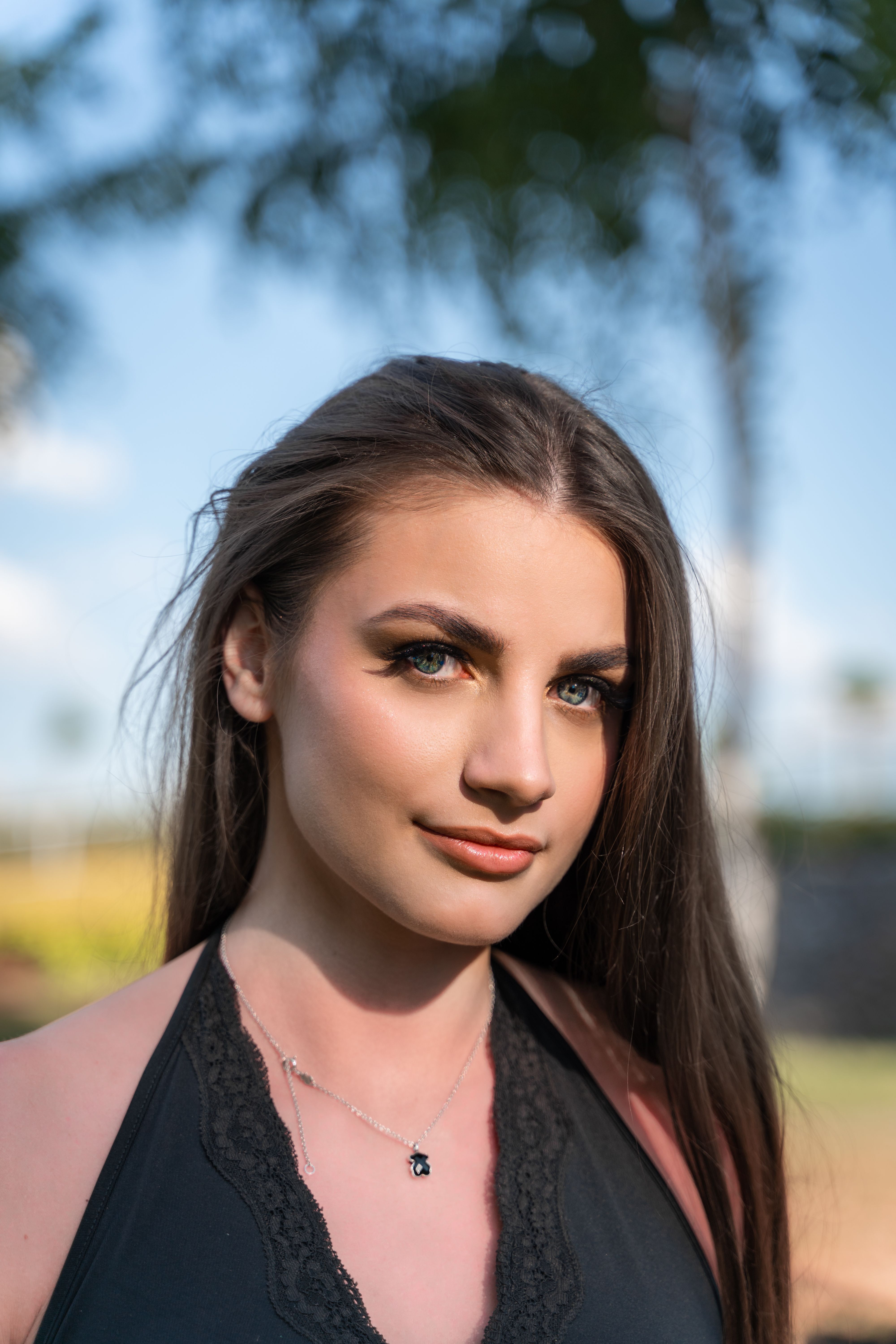 Close-up portrait of a young woman with long hair and a necklace, captured outdoors in natural light.