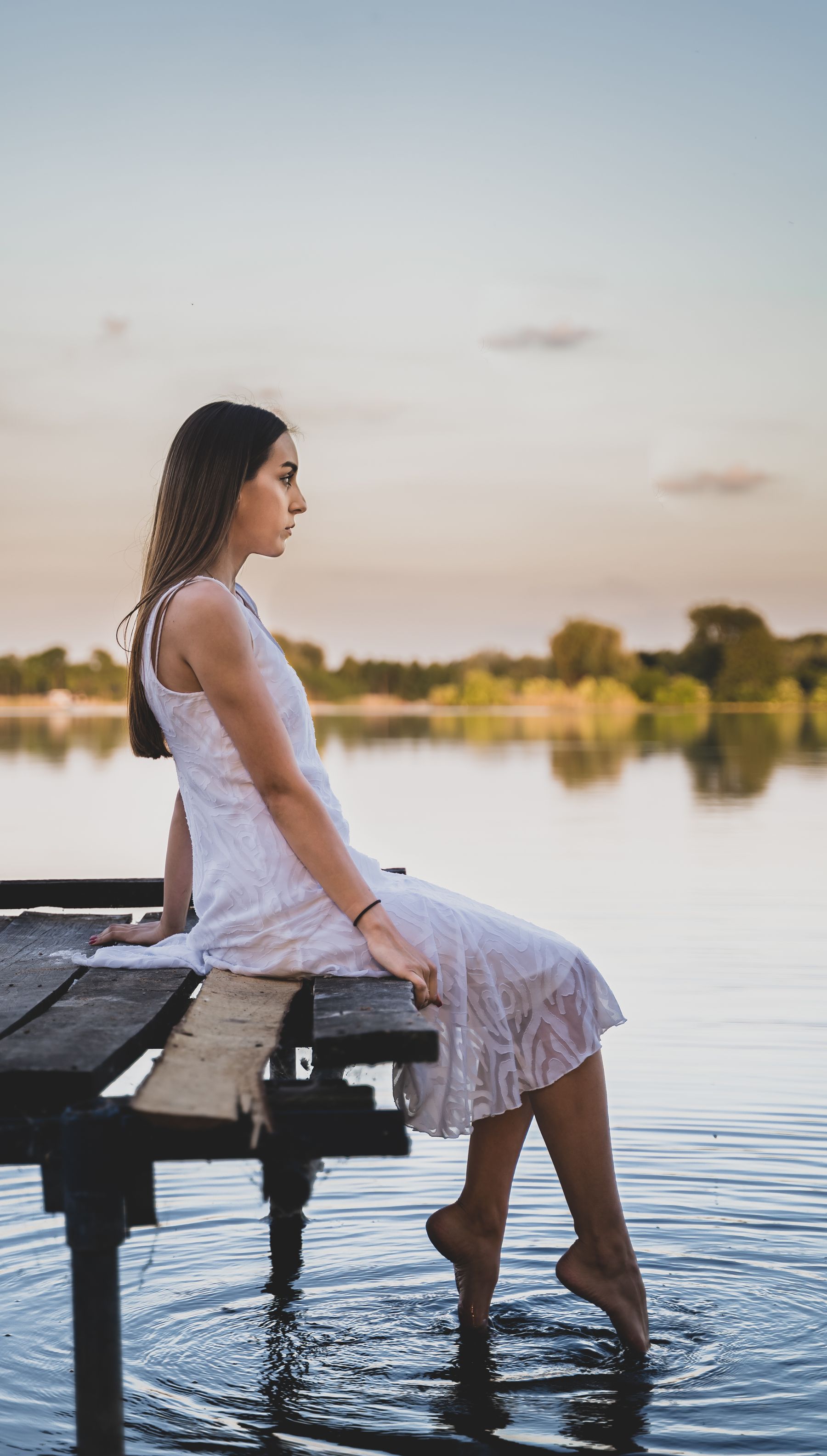 A woman in a white summer dress sitting on an old wooden dock with her feet in a calm lake, soft evening light over the water