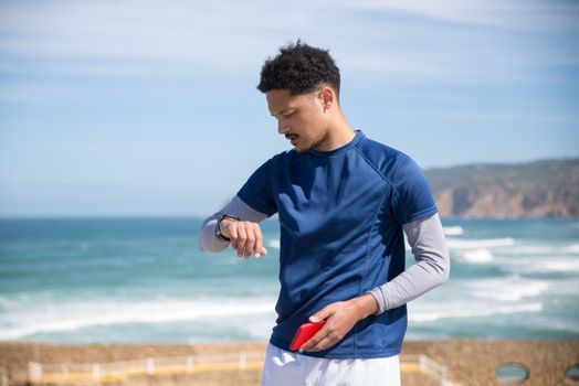 A sporty man checks his watch while standing by the ocean on a sunny day in Portugal.