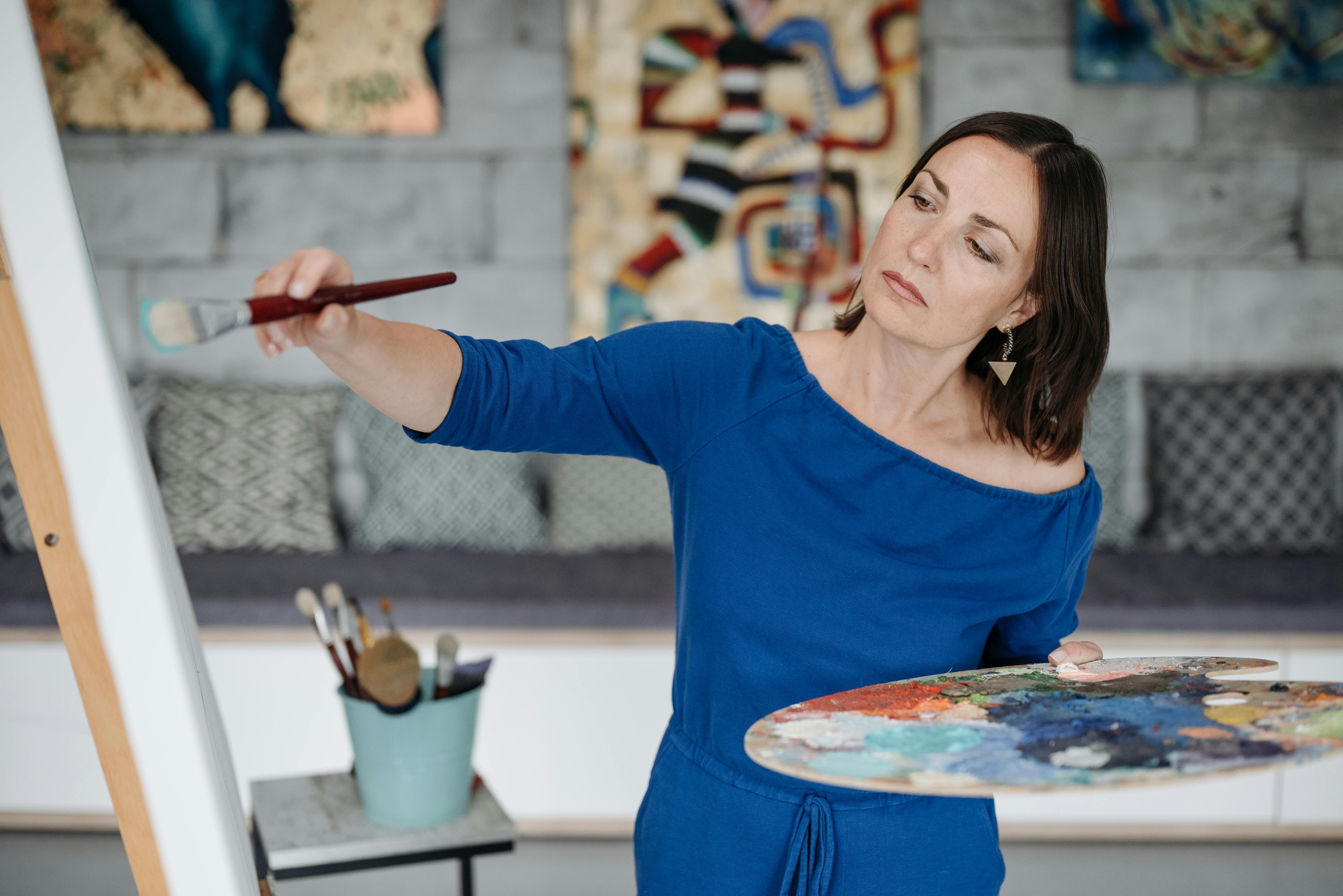 A woman artist intensely working on her painting indoors with a brush and paint palette.