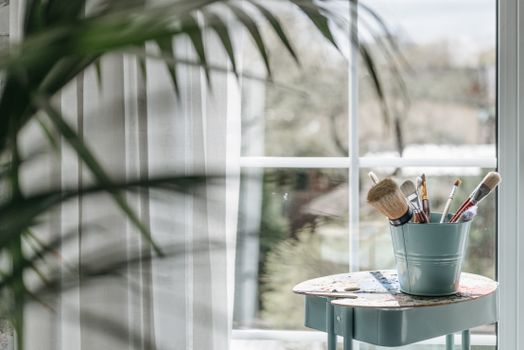 A bucket of art brushes on a table by a sunny window, inspiring creativity.