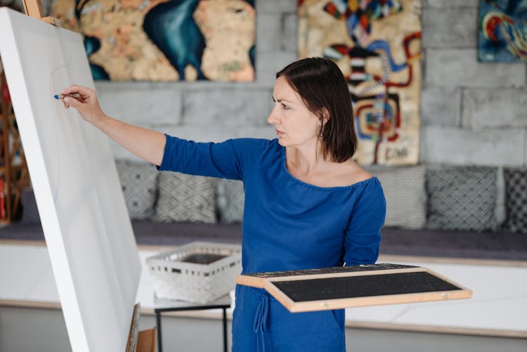 A Woman In Blue Long Sleeve Dress Drawing On White Canvass