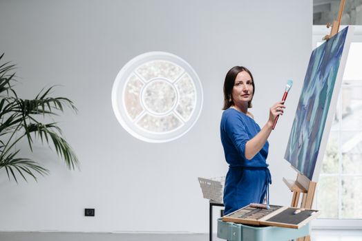 Woman artist in blue dress painting on canvas in well-lit studio with round window.