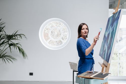 Free Woman artist in blue dress painting on canvas in well-lit studio with round window. Stock Photo