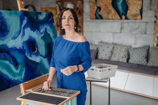 A female artist working on an abstract blue canvas using pastels in an art studio.