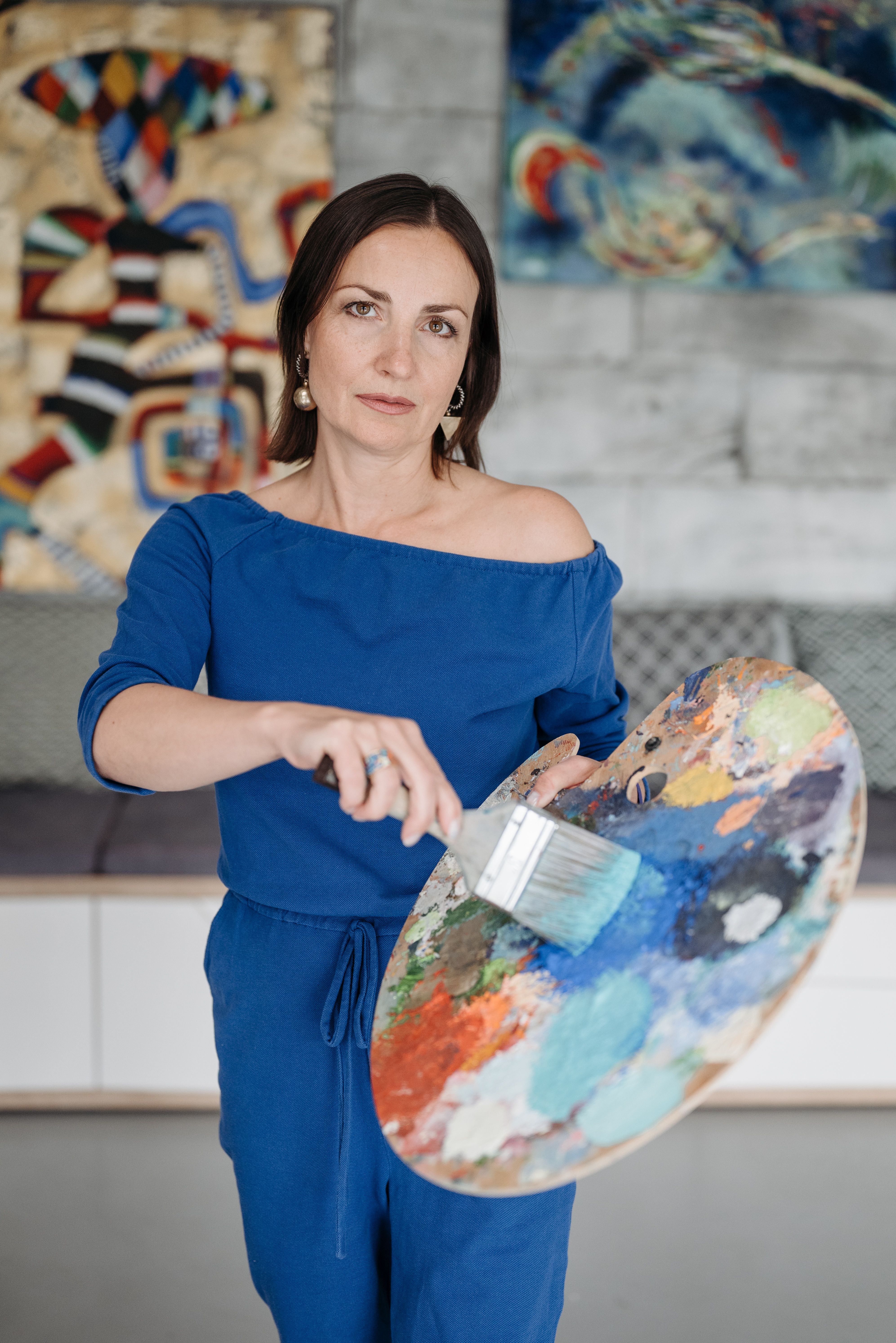 A woman artist in a blue outfit holding a paintbrush and colorful palette indoors.