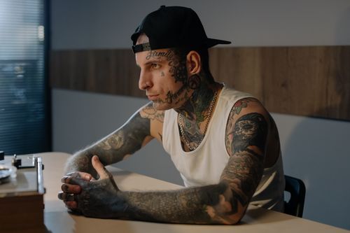 Free Side view of a tattooed man wearing a tank top and cap, sitting in a contemplative pose. Stock Photo