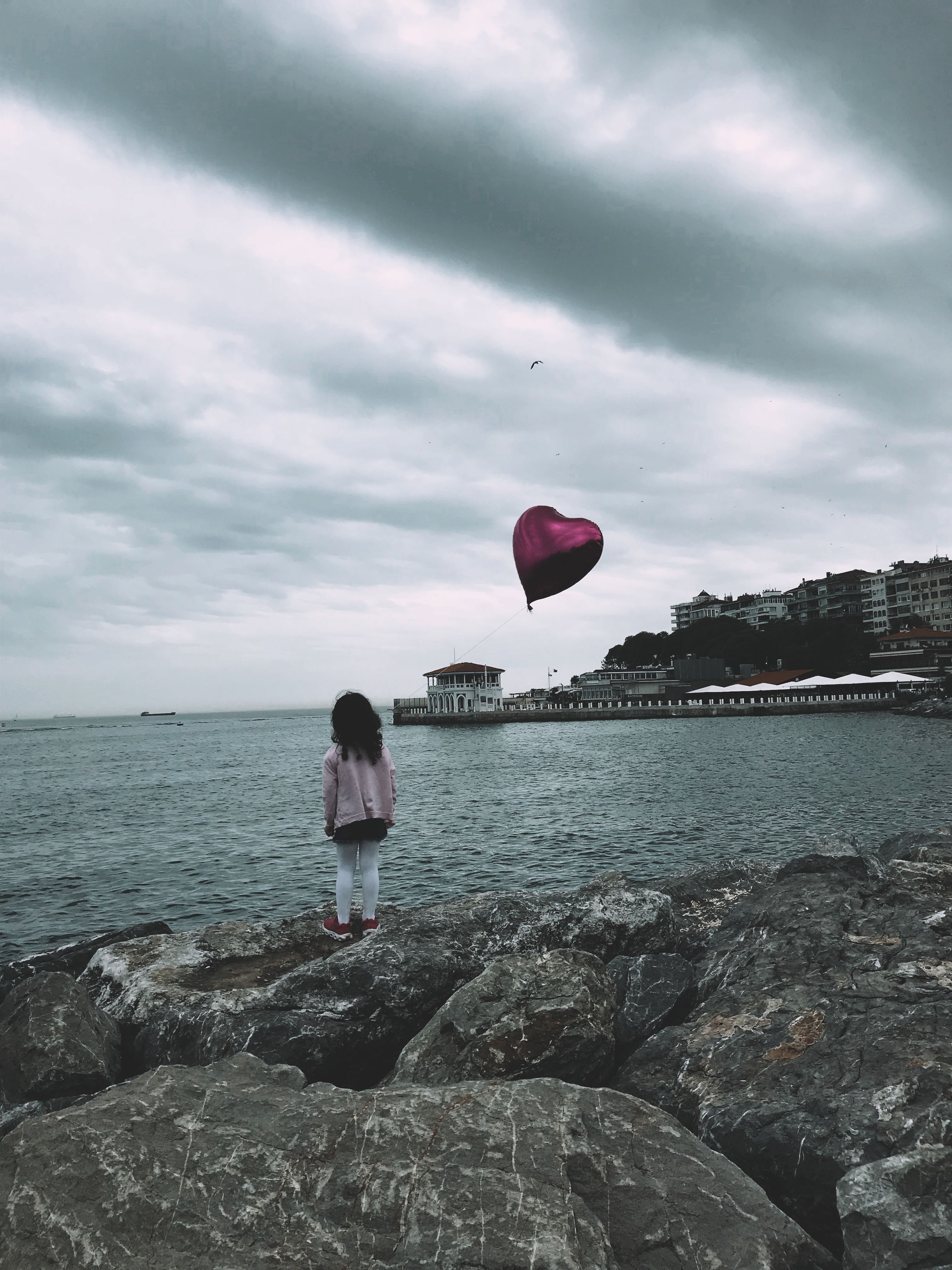 A child with a red heart balloon overlooking the sea on a cloudy day in Kadıköy, Istanbul.