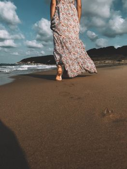 A woman in a floral dress walks barefoot along Şile Beach, Turkey, creating footprints in the sand under a partly cloudy sky.