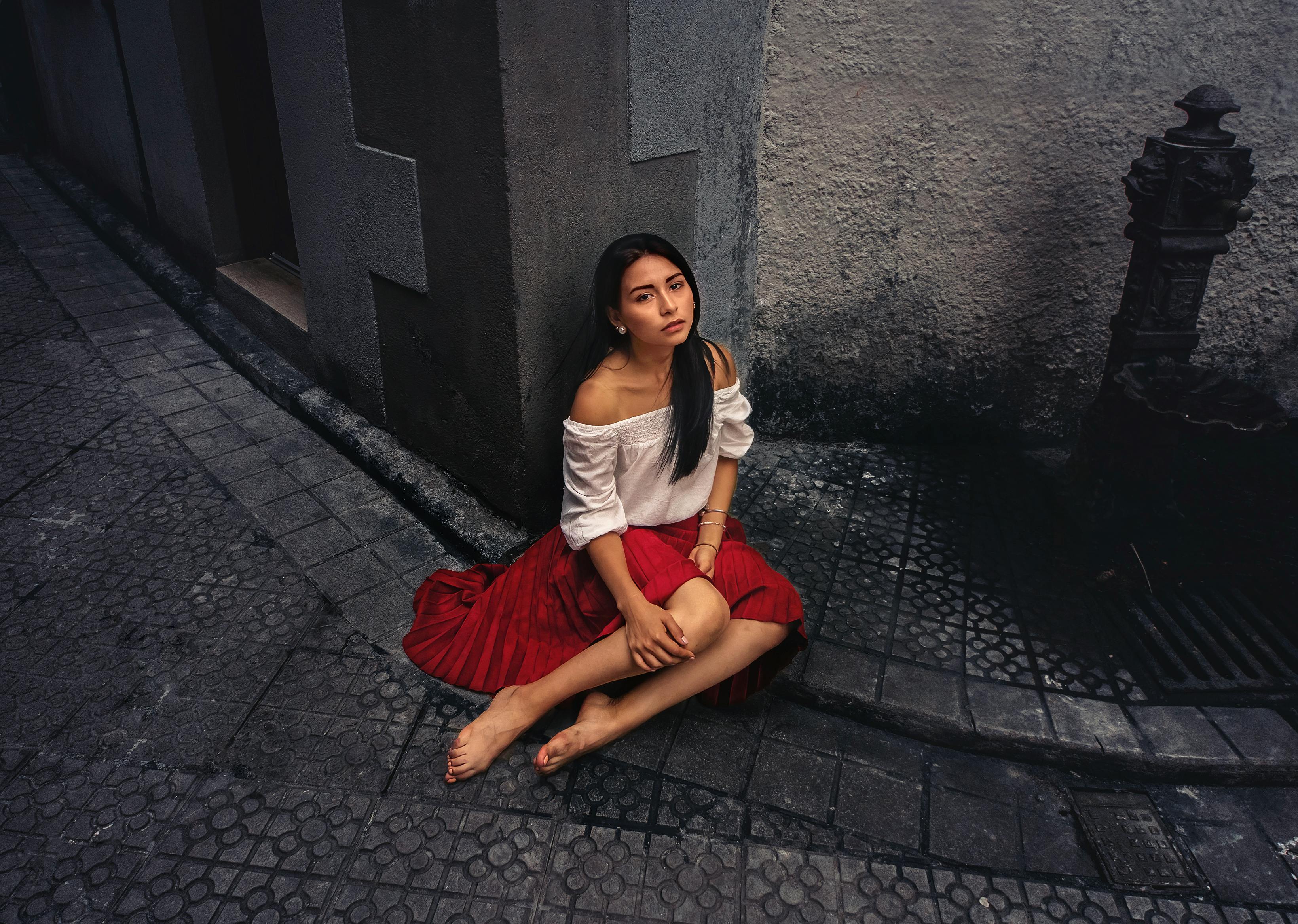 Free A contemplative woman in a red skirt sits barefoot on a city sidewalk, creating a striking urban portrait. Stock Photo