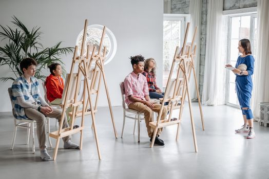 Students in an art class with easels, guided by a teacher in a bright studio setting.