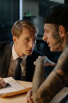 A detective interrogates a tattooed suspect in an office setting, highlighting tension.