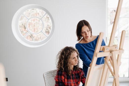 A teacher instructing a young student in an art class, focusing on painting techniques at an easel.