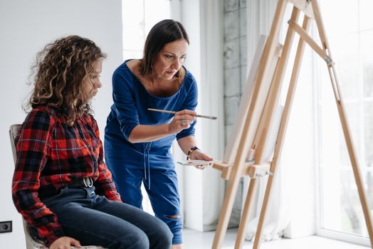 A teacher guides a student during an art class, demonstrating painting techniques on a canvas.