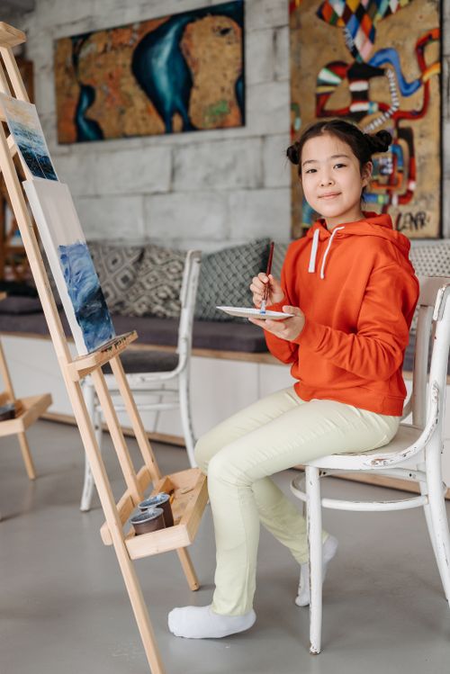 Free Teenage girl painting in a studio, wearing an orange hoodie and yellow pants, smiling and relaxed. Stock Photo
